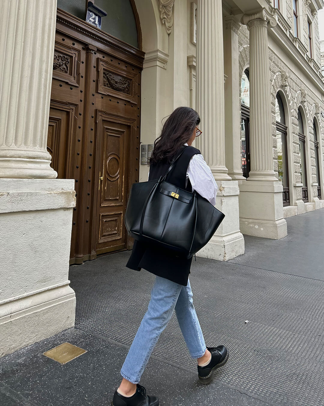 Black leather tote bag on shoulder, model walking outdoors, back view