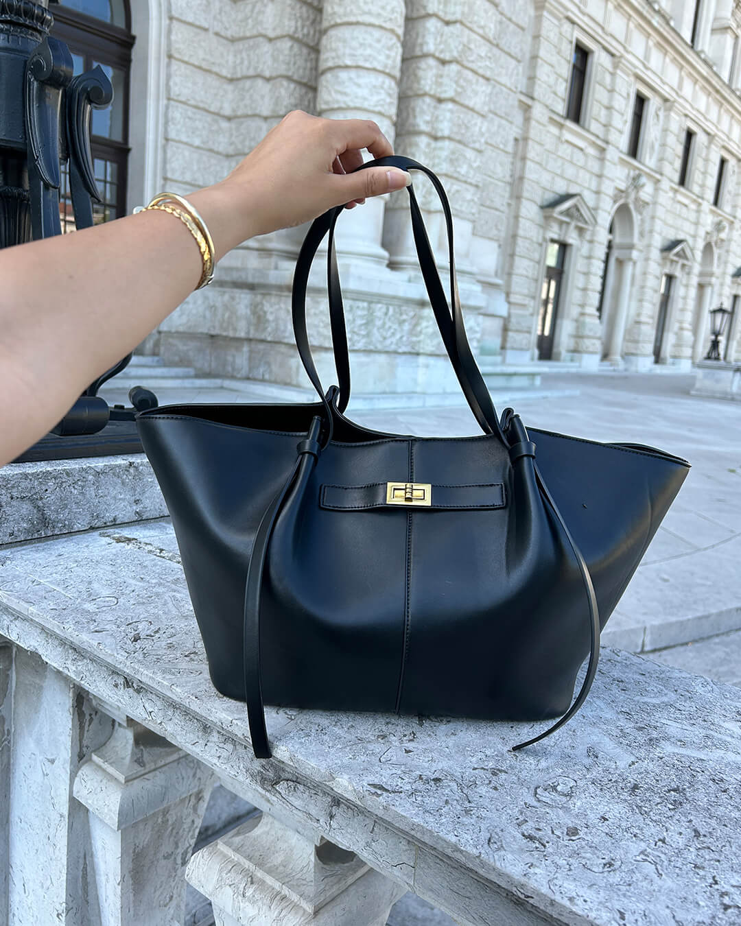 Black leather tote bag held by handles, outdoors on stone ledge