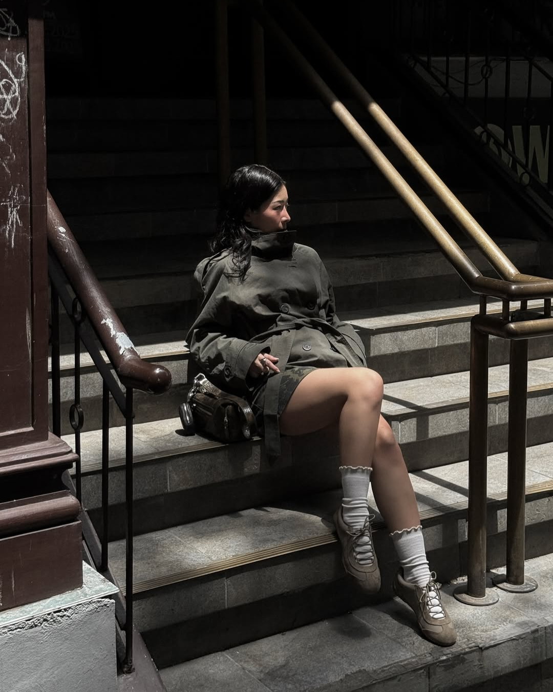 Olive green short belted trench coat, model seated on steps