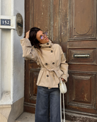 Beige short belted trench coat, model by a wooden door
