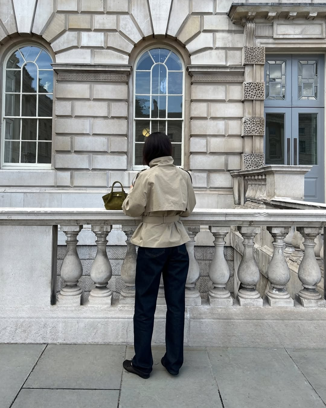 Beige short belted trench coat, back view leaning on railing