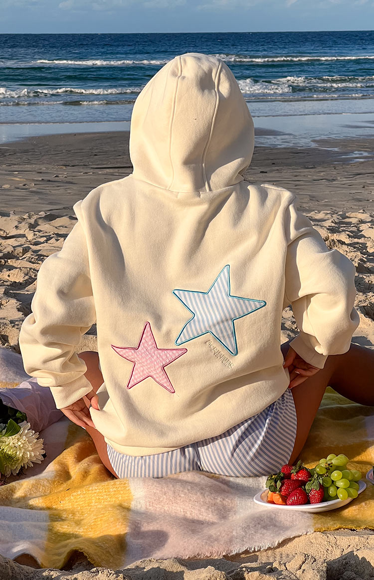 Cream hoodie with star patches, back view, model seated on beach